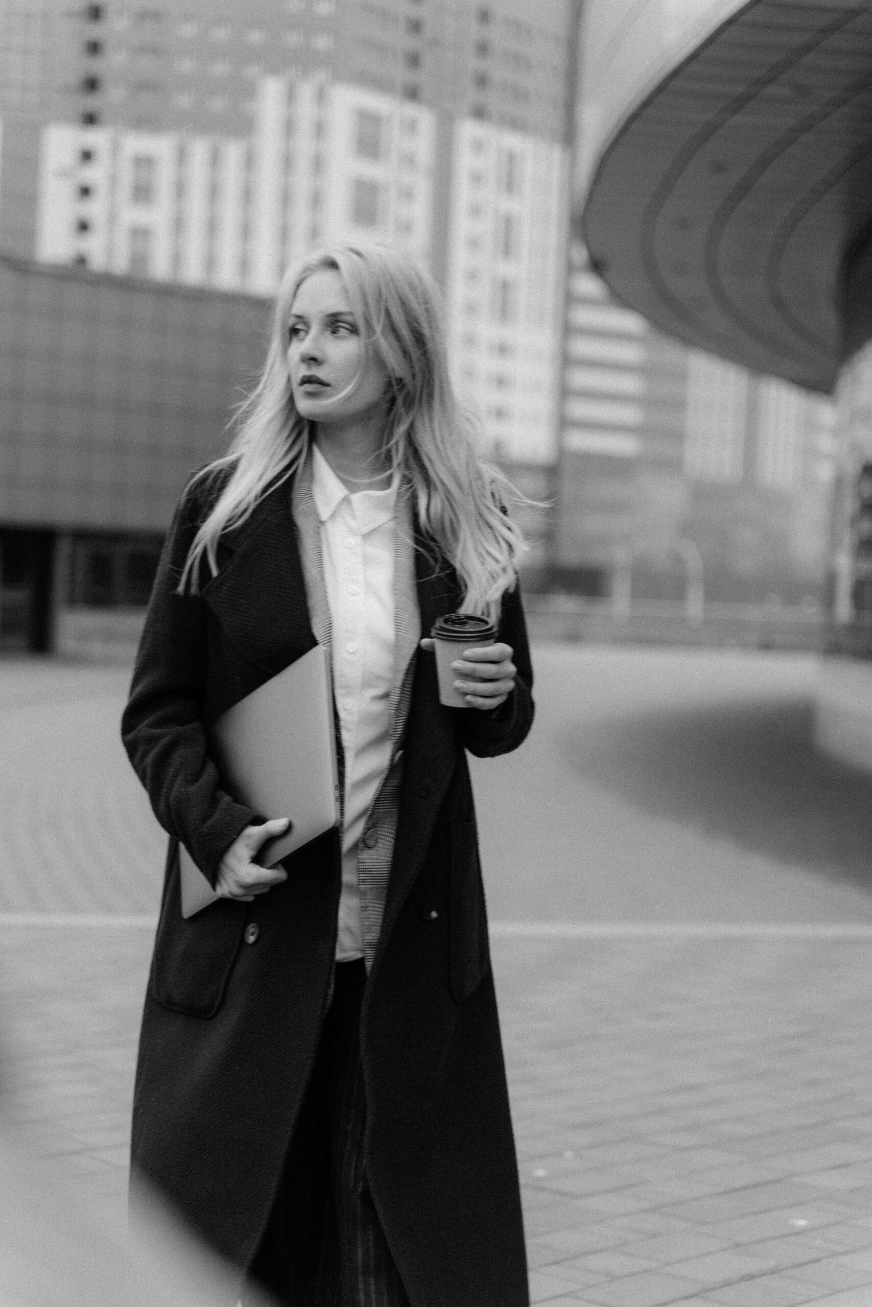 Elegant businesswoman walking in city holding a laptop and coffee, embodying leadership and style.